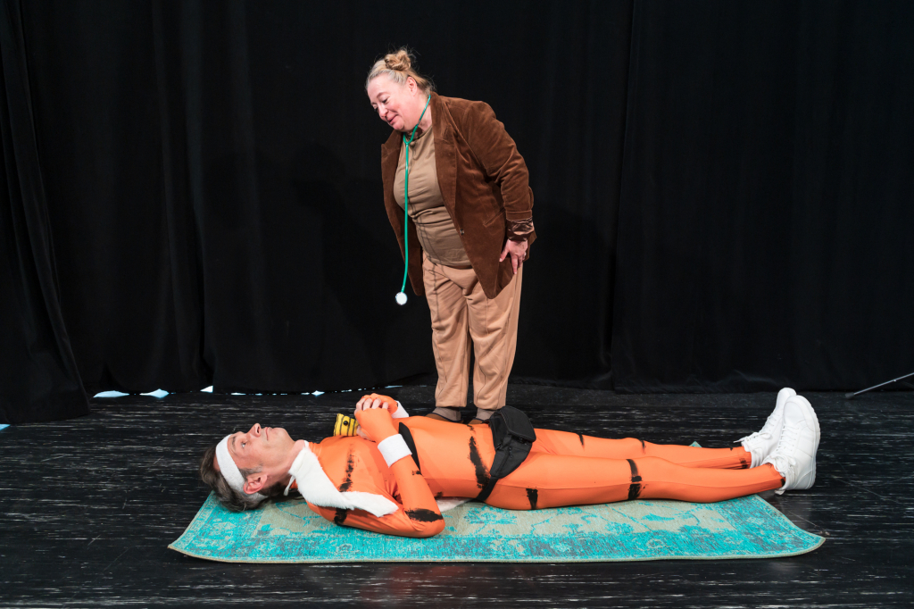 Person in braunem Anzug beugt sich mit grünem Stethoskop über eine auf einer blauen Matte liegende Person im orangefarbenen Ganzkörperanzug mit weißen Schuhen.