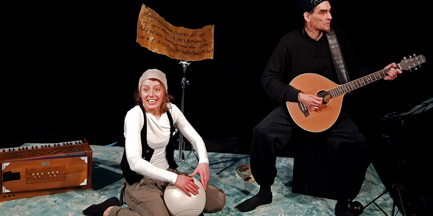 Zwei Personen auf einer Bühne, eine sitzt auf dem Boden und spielt eine Handtrommel, die andere steht und spielt Gitarre, im Hintergrund ein Harmonium und ein Schild mit handgeschriebenem Text.