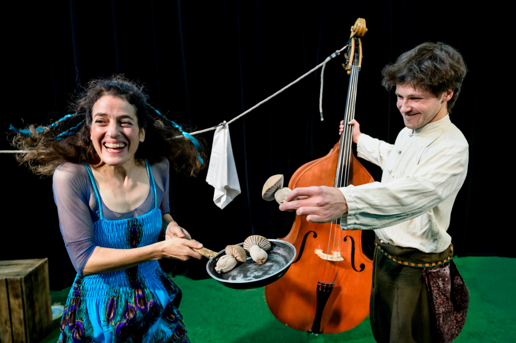 Frau in blauem Kleid hält Pfanne mit Pilzen, Mann in weißem Hemd hält Kontrabass und eine Muschel, schwarze Bühne mit grünem Boden.