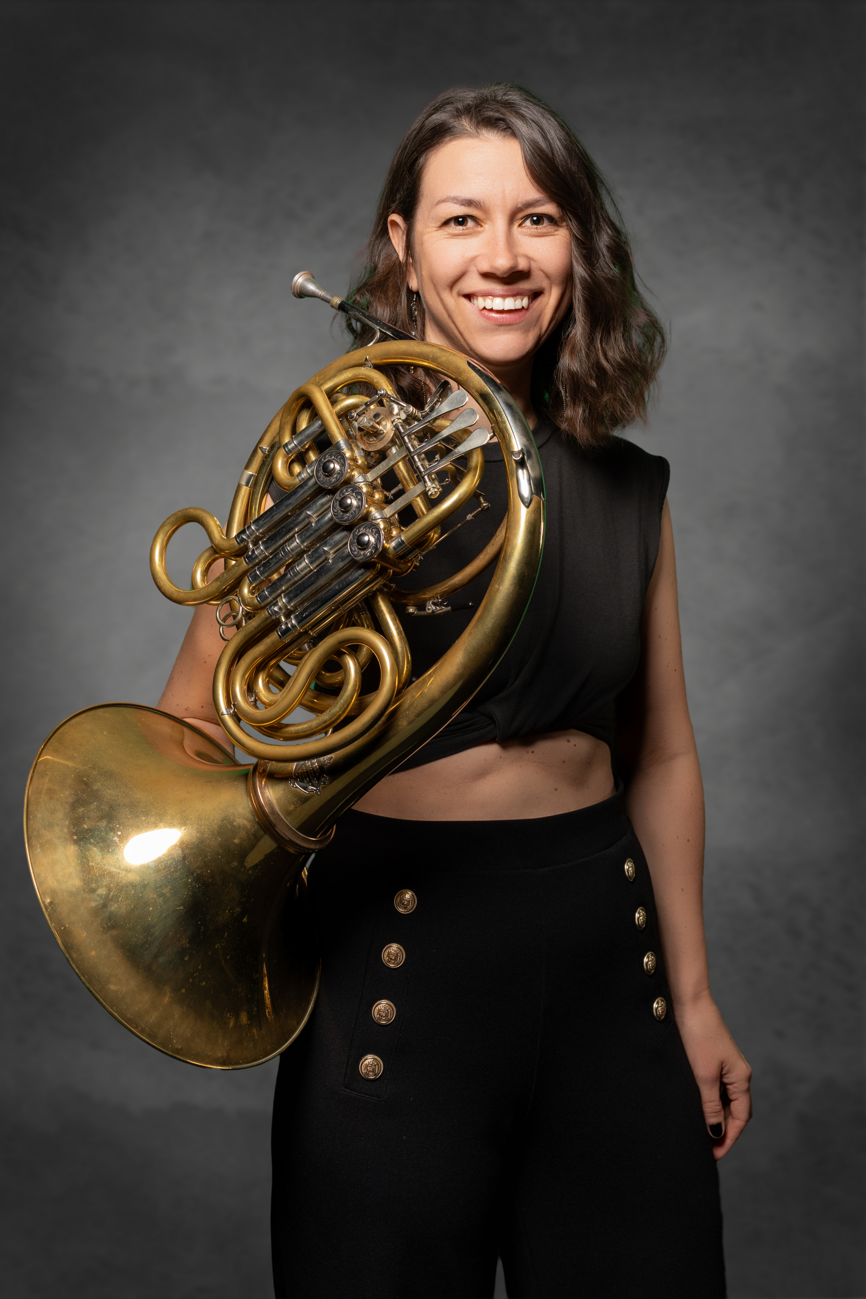 Junge Frau mit einer Konzert-Horn in der Hand, lächelnd vor einem grauen Hintergrund.