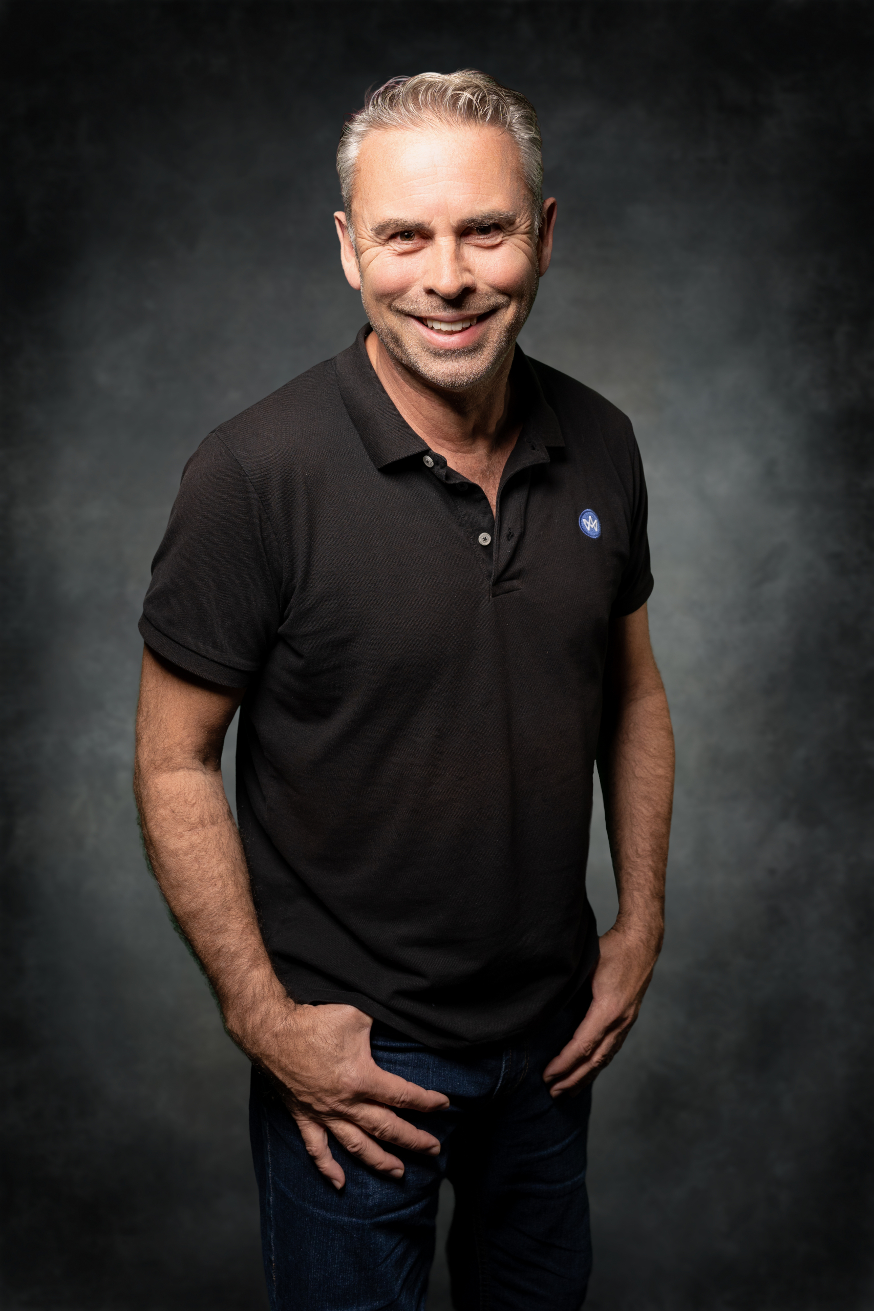 Lächelnder Mann in schwarzem Poloshirt, steht aufrecht mit den Händen in den Hosentaschen.