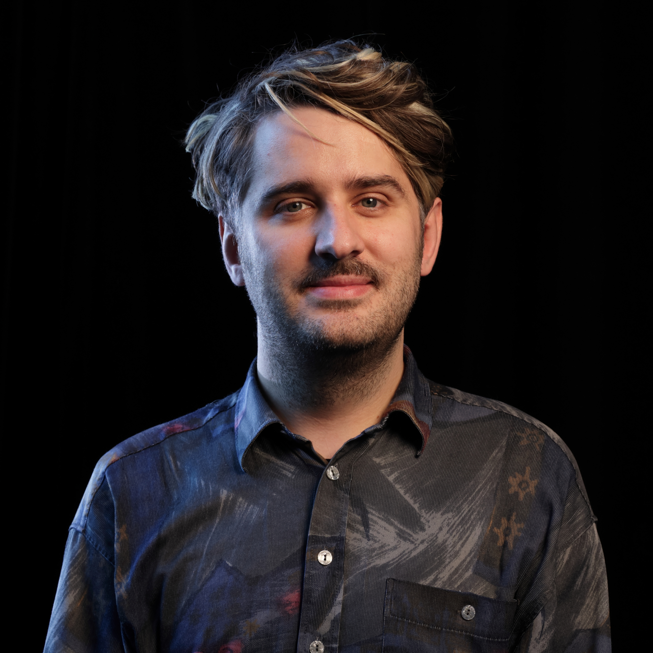 Oberkörper eines Mannes mit kurzem, gewelltem Haar, farbig gemustertem Hemd bvor schwarzem Hintergrund.