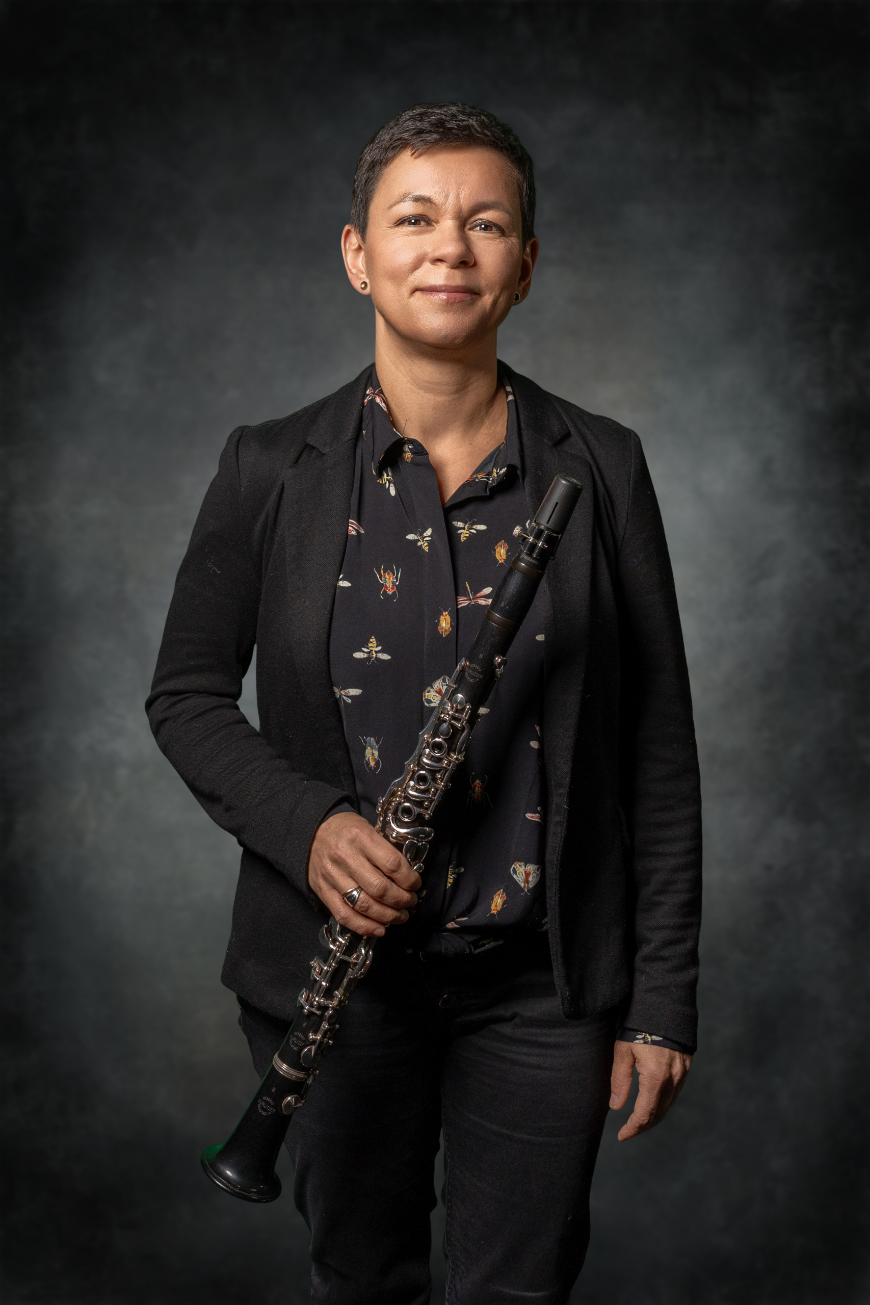Porträt einer Musikerin mit Klarinette, in schwarzem Blazer und bedrucktem Hemd, vor dunklem Hintergrund.