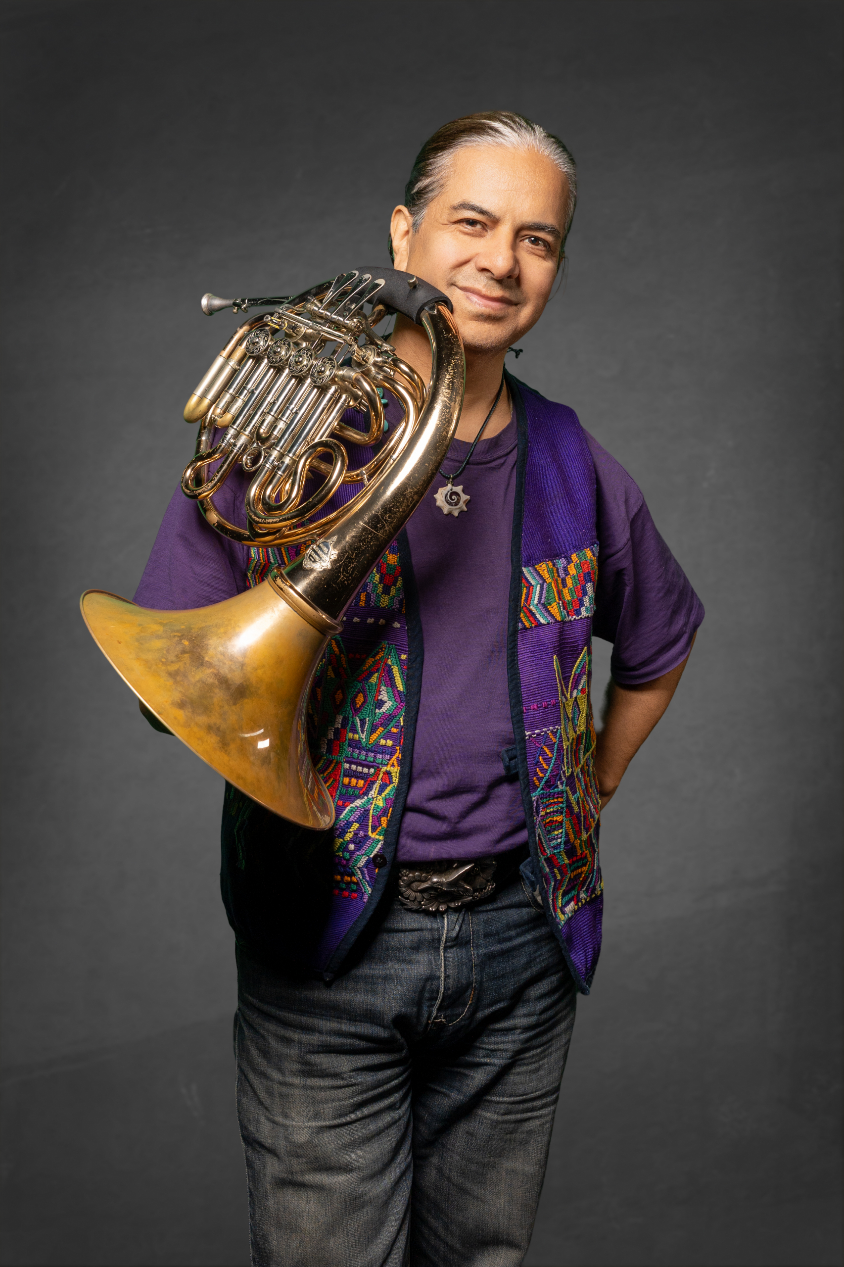 Mann mit einem französischen Horn, trägt bunte Weste und lächelt vor einem grauen Hintergrund.