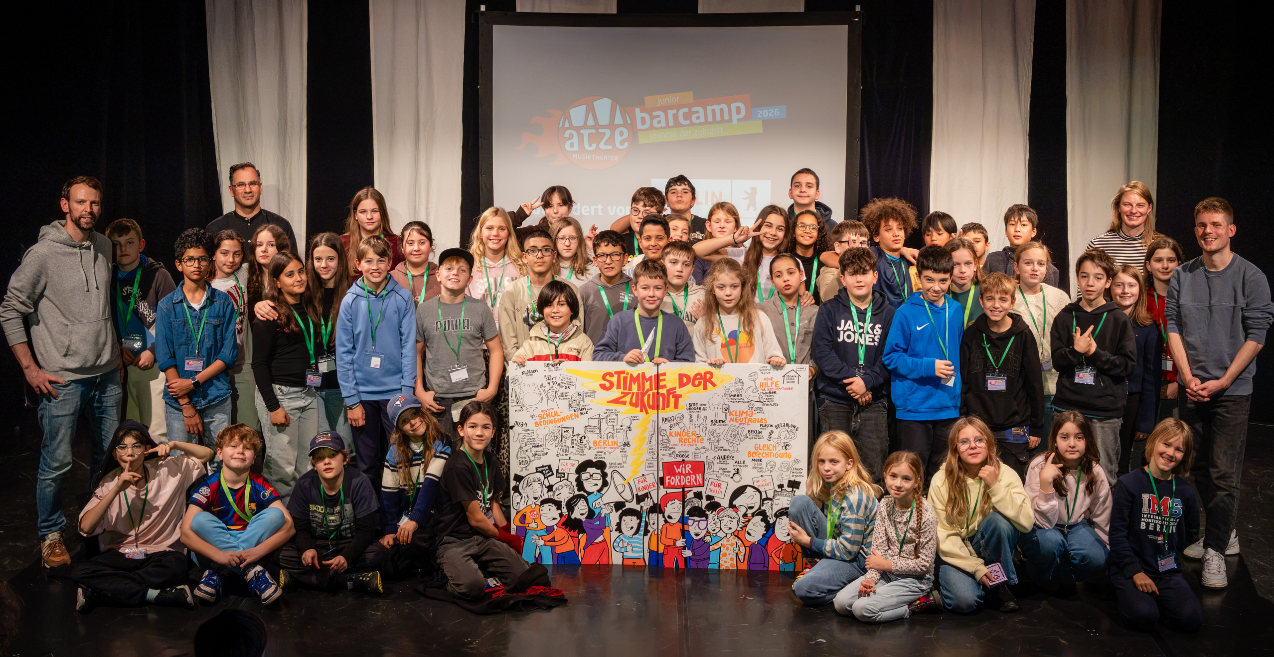 Große Gruppe von Kindern und Jugendlichen mit einigen Erwachsenen auf einer Bühne vor einer Leinwand mit dem Schriftzug 'atze barcamp'