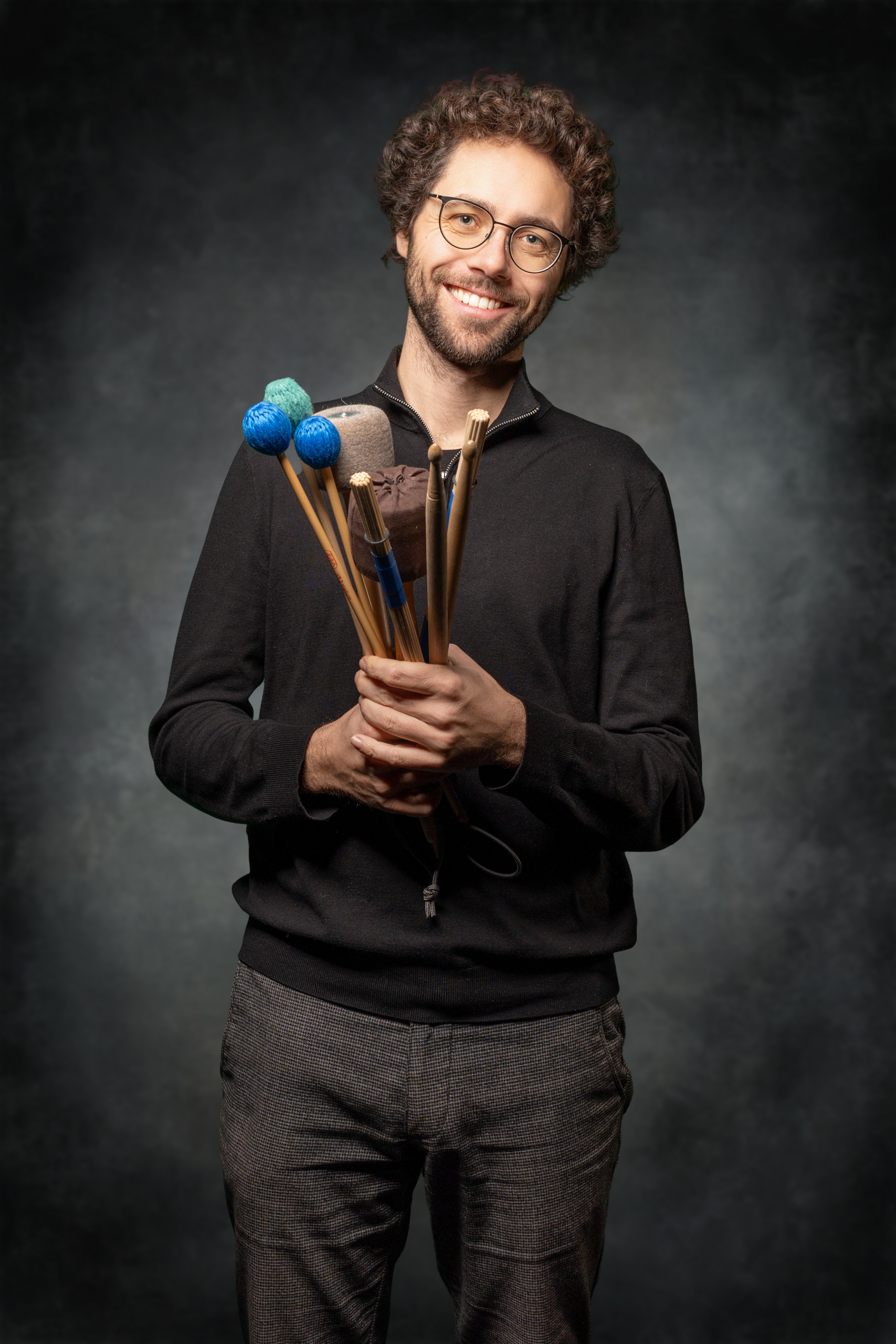 Mann hält verschiedene Schlägel in der Hand, lächelt freundlich in einem dunklen Hintergrund.