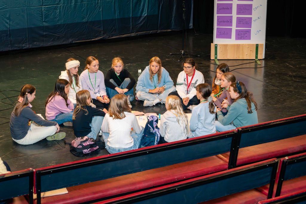 Gruppe von Jugendlichen sitzt im Kreis auf dem Boden einer Bühne, im Hintergrund ein Flipchart mit Notizzetteln