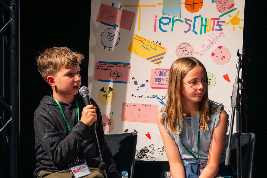 Zwei Kinder mit Namensschildern sitzen vor einem Plakat mit bunten Zeichnungen und dem Schriftzug 'Tierschutz'