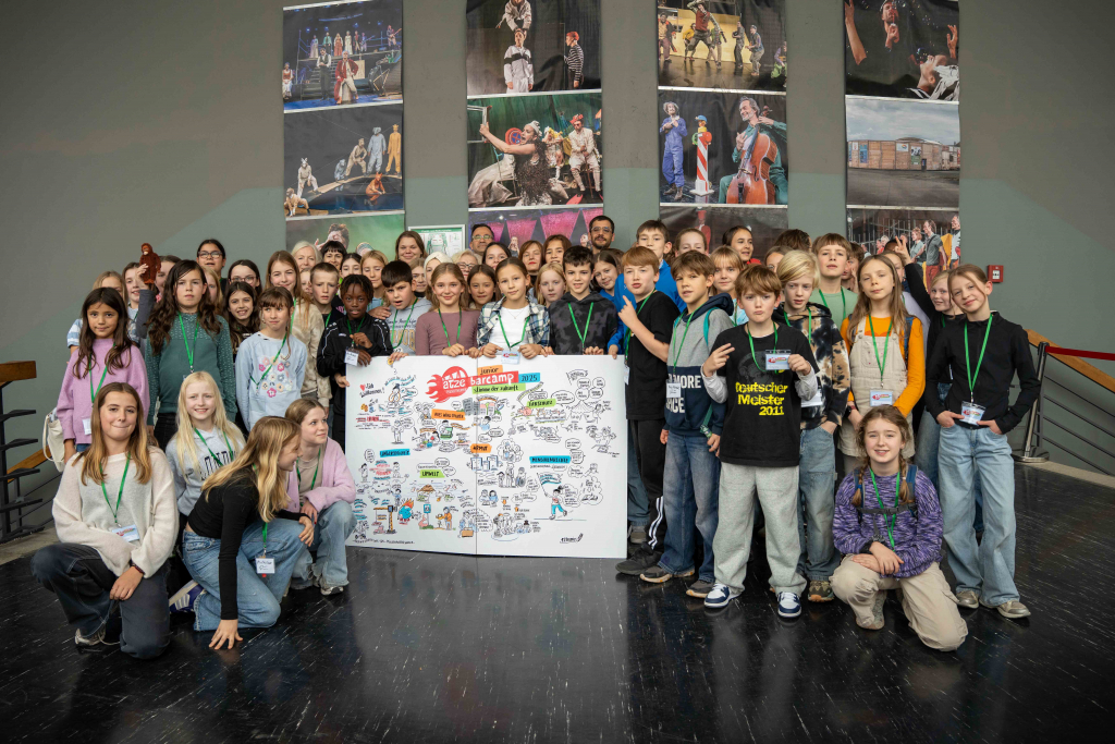 Große Gruppe von Kindern und Jugendlichen mit Namensschildern steht und kniet vor einer großen gezeichneten Plakatwand in einem Raum mit Bühnenbildern an der Wand