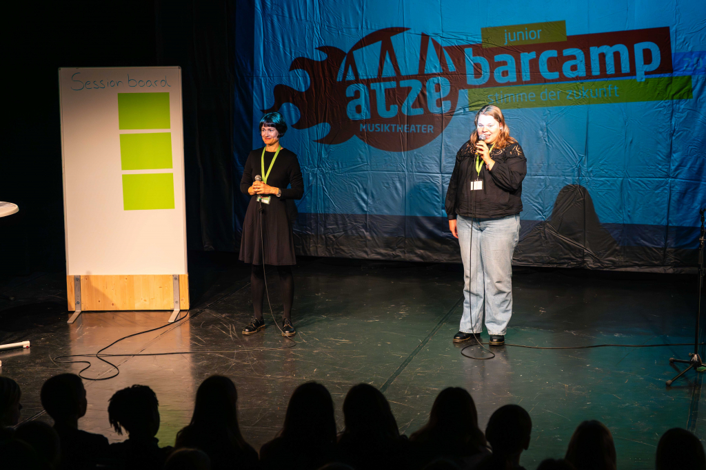 Zwei Frauen stehen auf einer Bühne vor einer Leinwand mit dem Schriftzug 'atze junior barcamp' und einem Session-Board mit drei grünen Zetteln