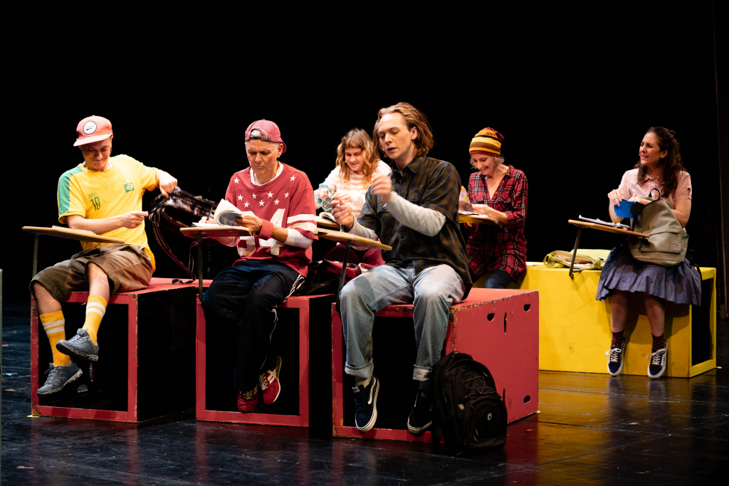 Fünf Personen sitzen auf bunten Holzkisten auf einer Bühne, einige halten Bücher oder Schreibutensilien, eine Person trägt eine gelbe Mütze.