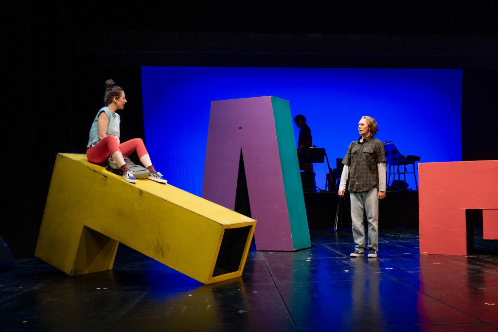 Zwei Personen auf Bühne mit bunten, geometrischen Bühnenobjekten vor blauem Hintergrund, eine sitzt auf gelbem Kasten, andere steht.