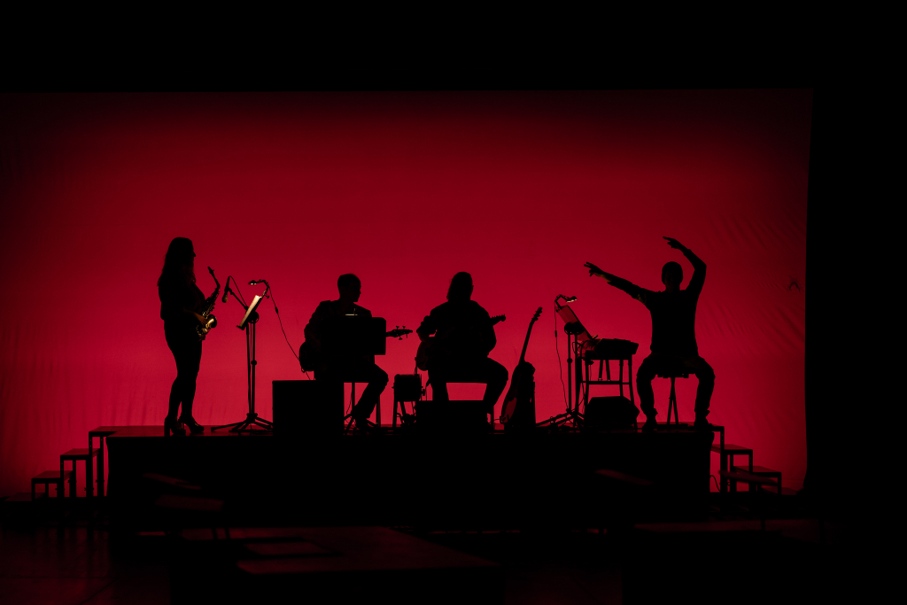 Vier Musiker als schwarze Silhouetten vor rotem Hintergrund, zwei sitzen, einer steht mit Saxofon, einer gestikuliert mit erhobenen Armen.