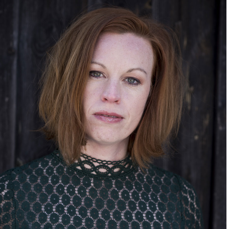 Portrait einer Frau mit kurzen, roten Haaren und einem grünen Spitzenoberteil, vor einer dunklen Holzoberfläche.