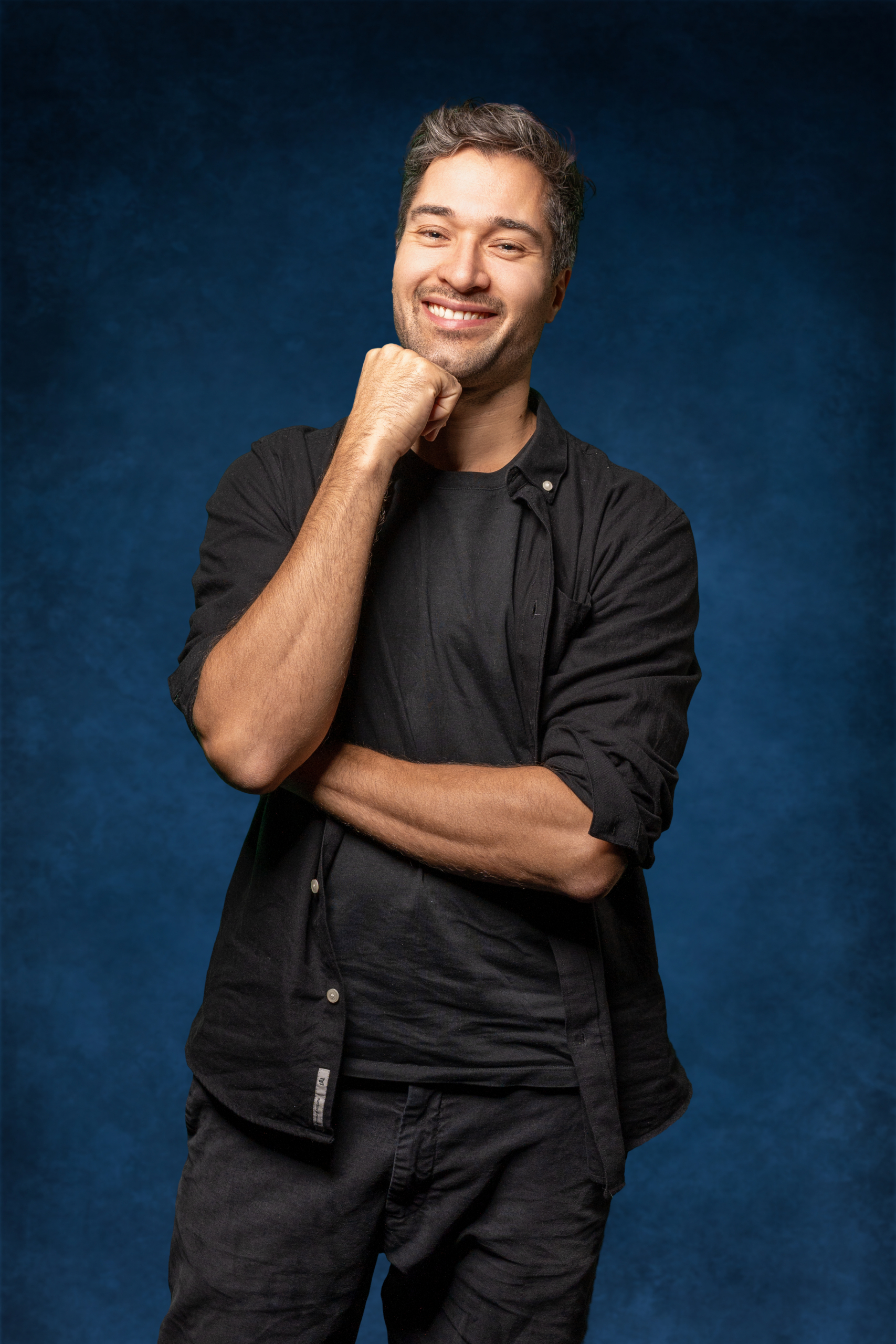 Lächelnder Mann in schwarzer Kleidung, mit gefalteten Armen vor einem blauen Hintergrund.