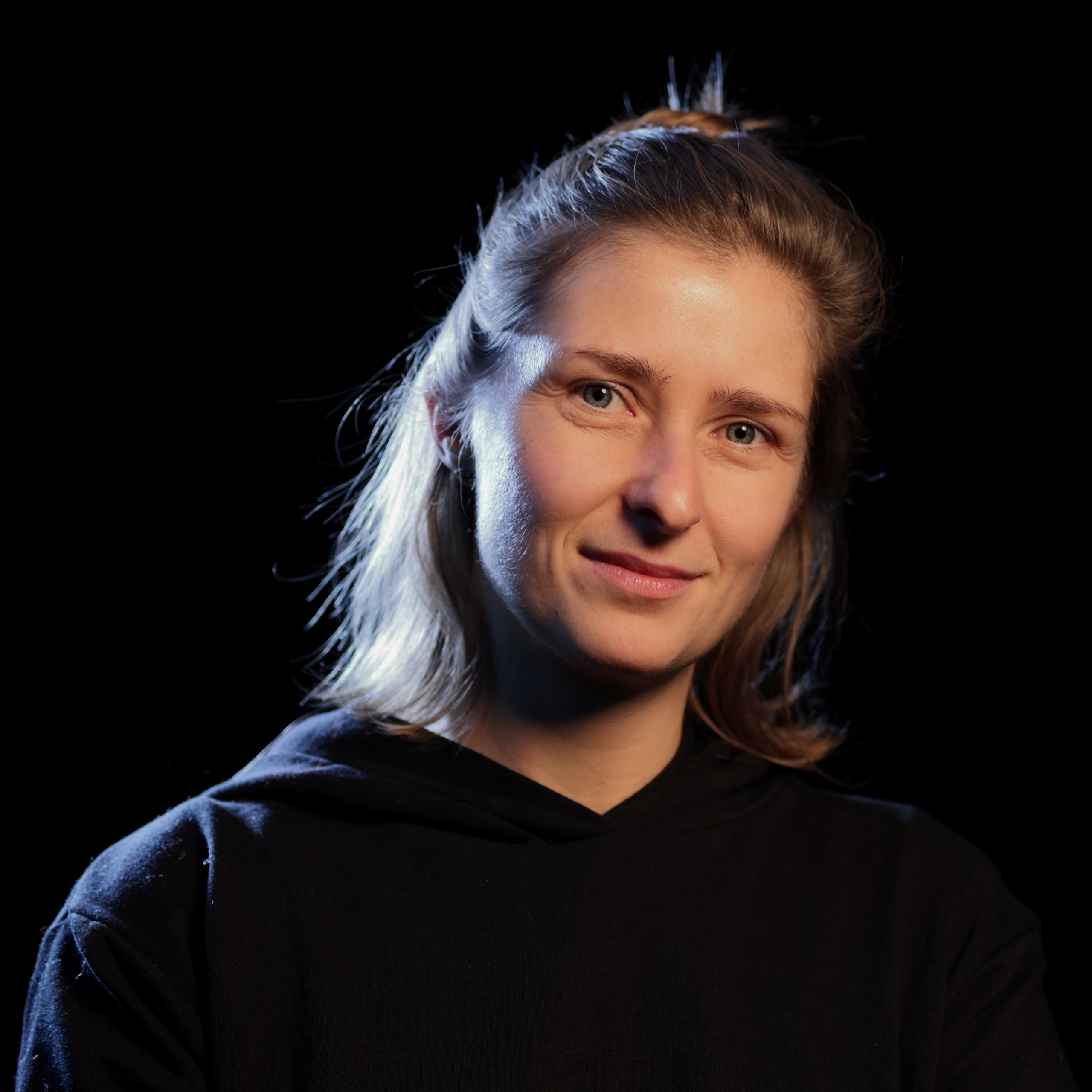 Porträt einer Frau mit hellen Haaren und einem dunklen Kapuzenpullover vor einem schwarzen Hintergrund.