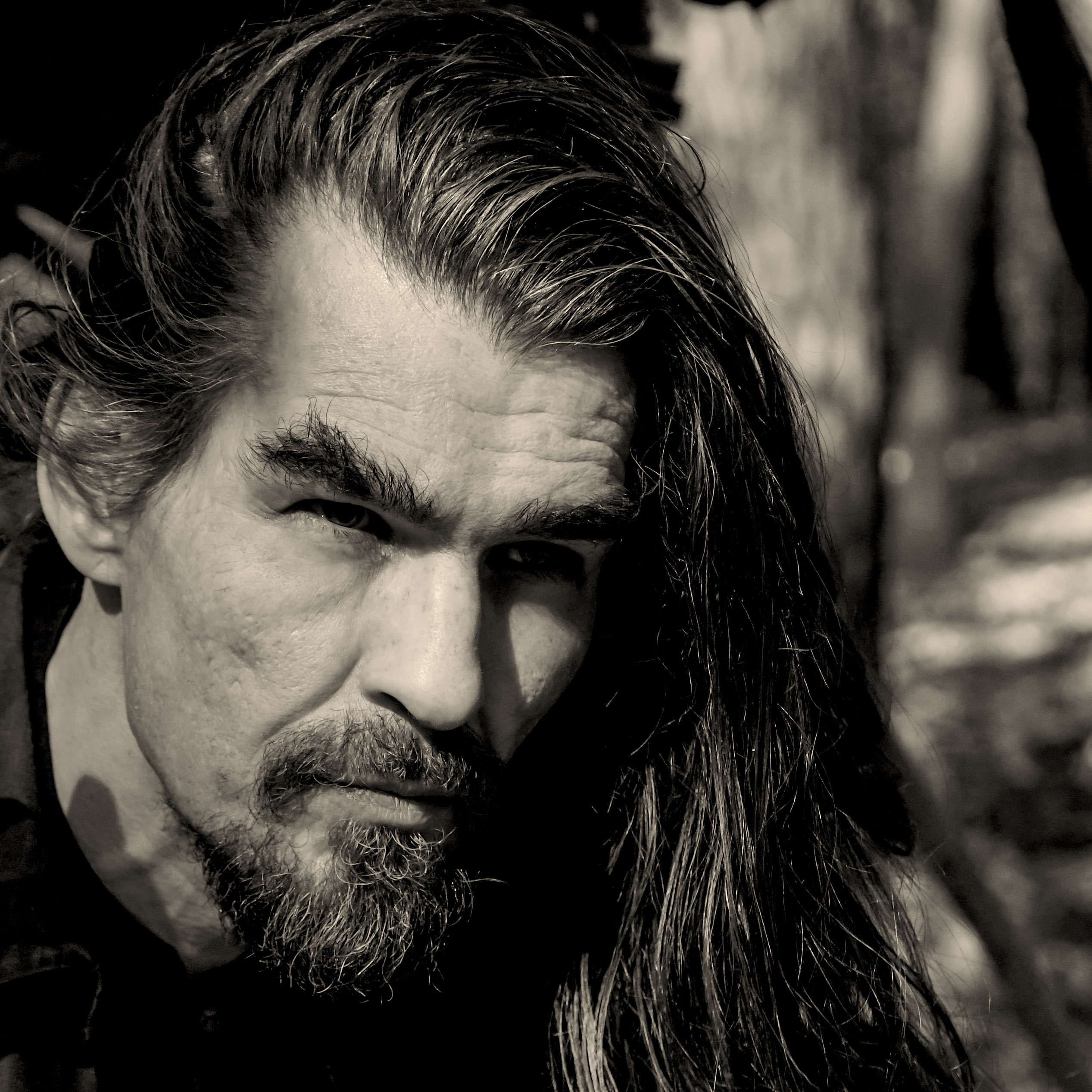 Schwarz-weiß Porträt eines Mannes mit langen Haaren und Bart, der ernst in die Kamera schaut.