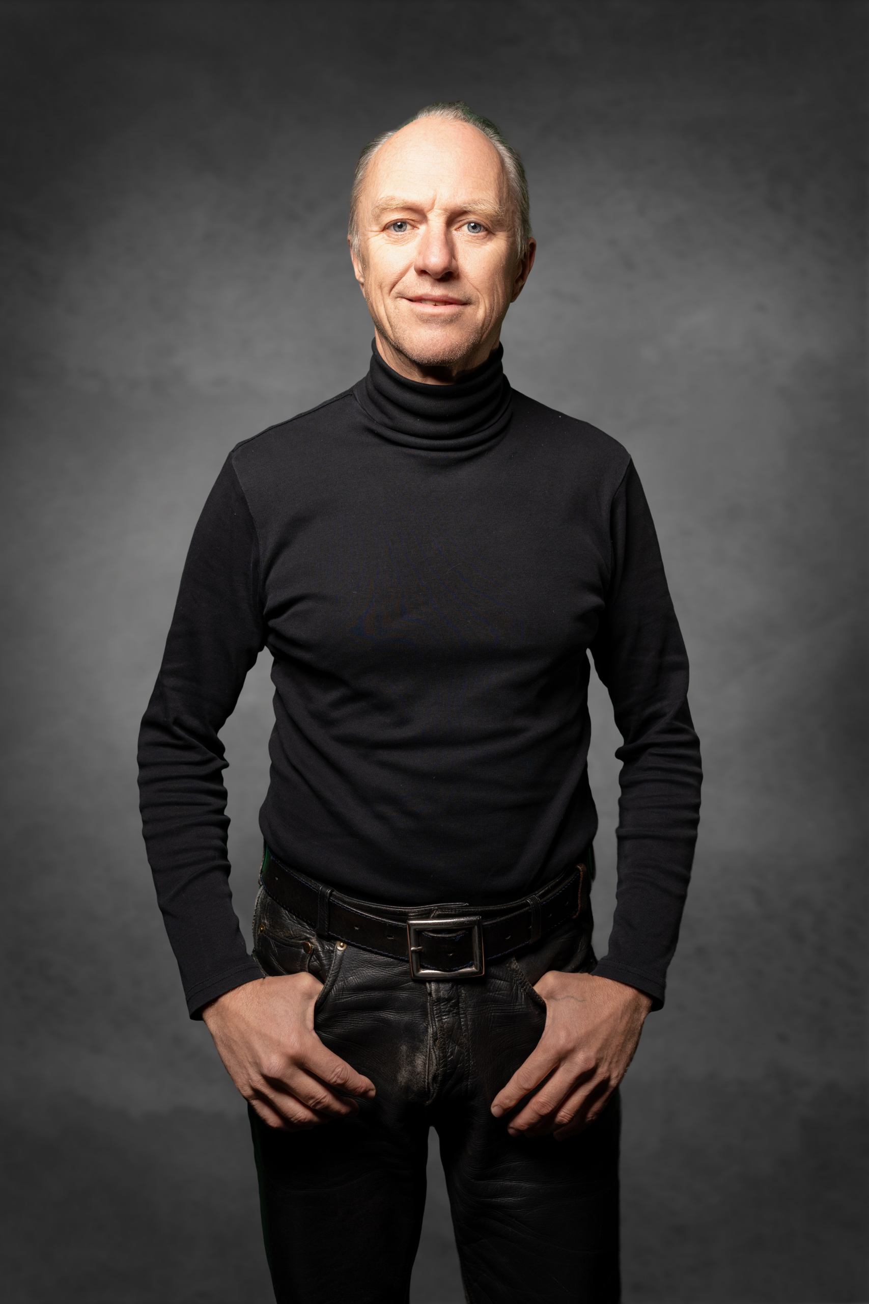 Mann im schwarzen Rollkragenpullover und Ledergürtel, lächelt für das Porträt auf einem grauen Hintergrund.