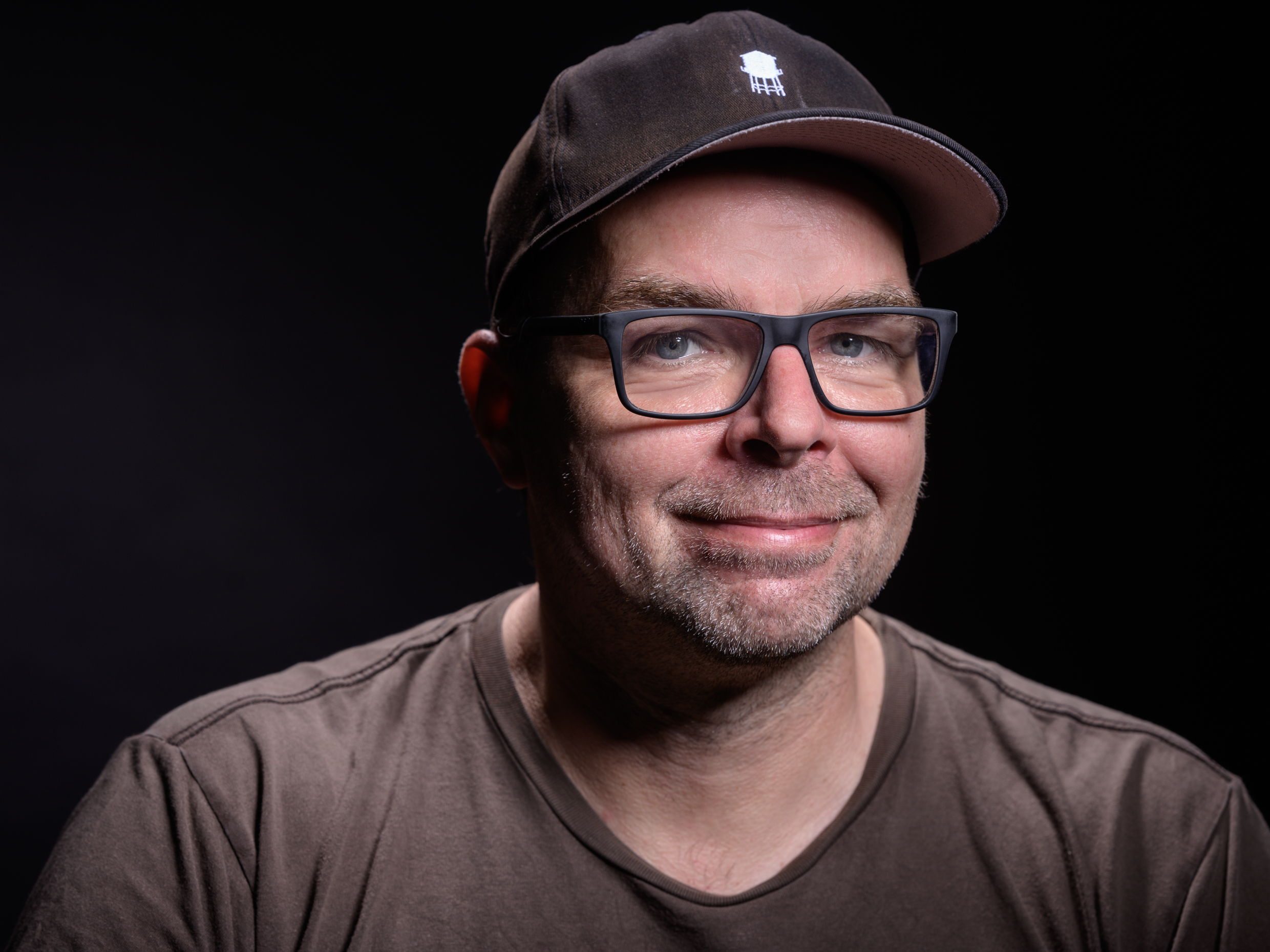 Mann mit Baseballkappe und Brille, trägt ein braunes T-Shirt, vor dunklem Hintergrund.