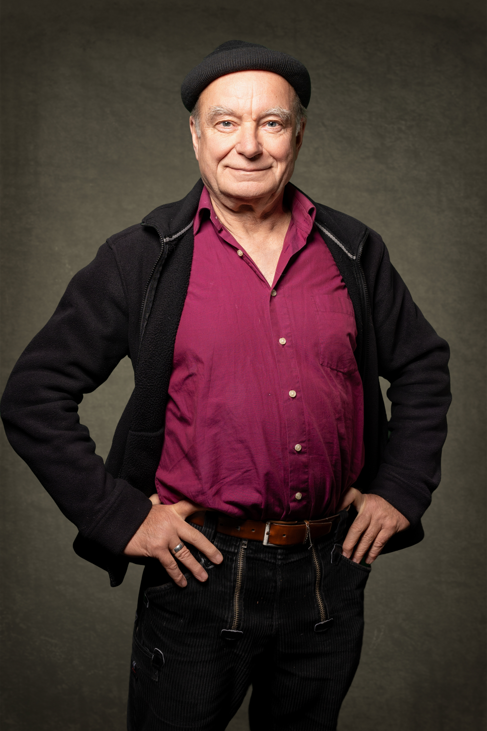 Porträt eines älteren Mannes mit Schmuddeljacke und schwarzer Mütze, der lächelt und die Hände in die Hüften gestemmt hat.