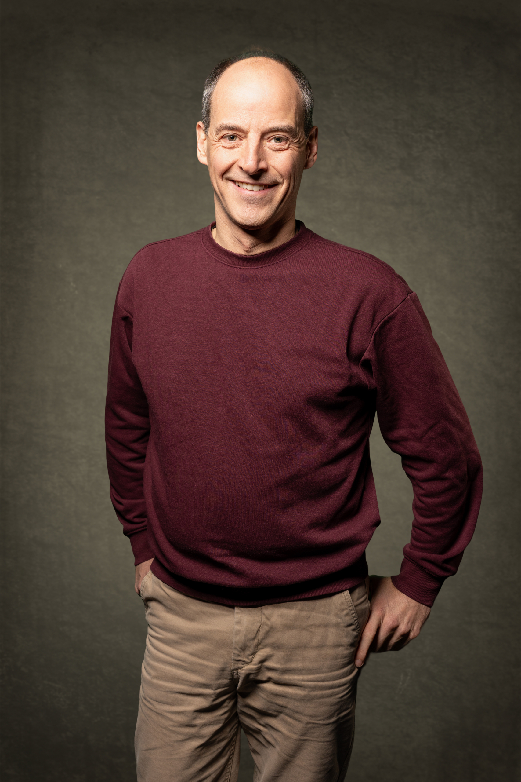 Mann in burgunderfarbenem Sweatshirt und hellen Hosen, lächelnd vor neutralem Hintergrund.