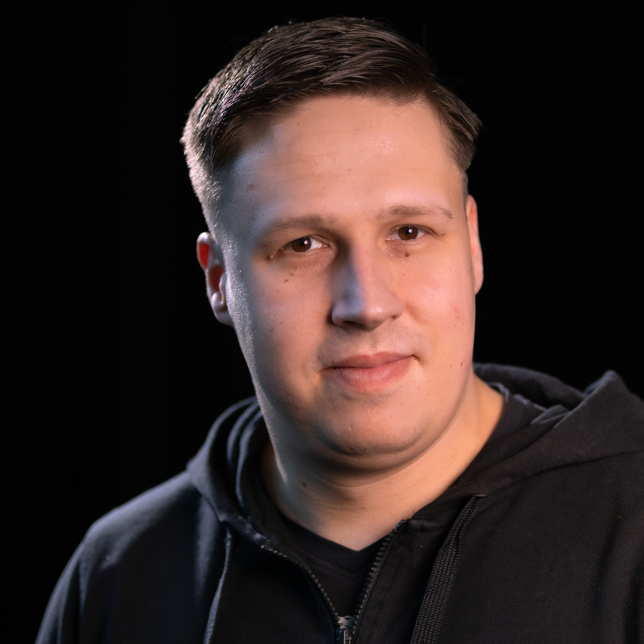 Mann mit kurzem, braunem Haar trägt schwarzen Kapuzenpullover vor schwarzem Hintergrund.