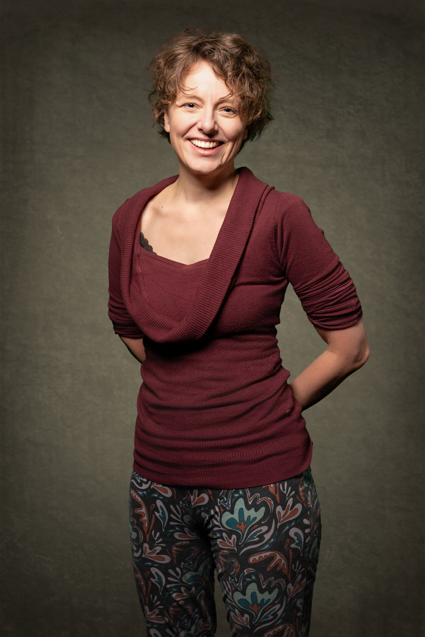 Frau mit lockigem Haar trägt ein burgunderfarbenes Oberteil und gemusterte Hose, lächelt vor neutralem Hintergrund.