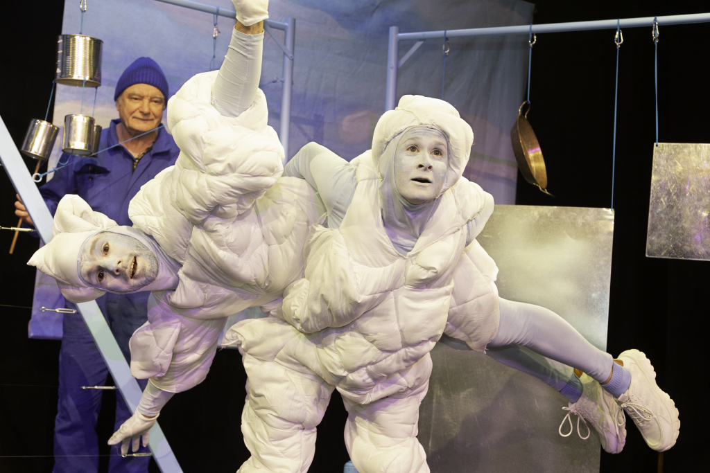 Zwei Personen in weißen, gepolsterten Anzügen posieren mit ausgestreckten Armen und Beinen, im Hintergrund hängt Küchenutensilien und eine dritte Person in blauer Kleidung.