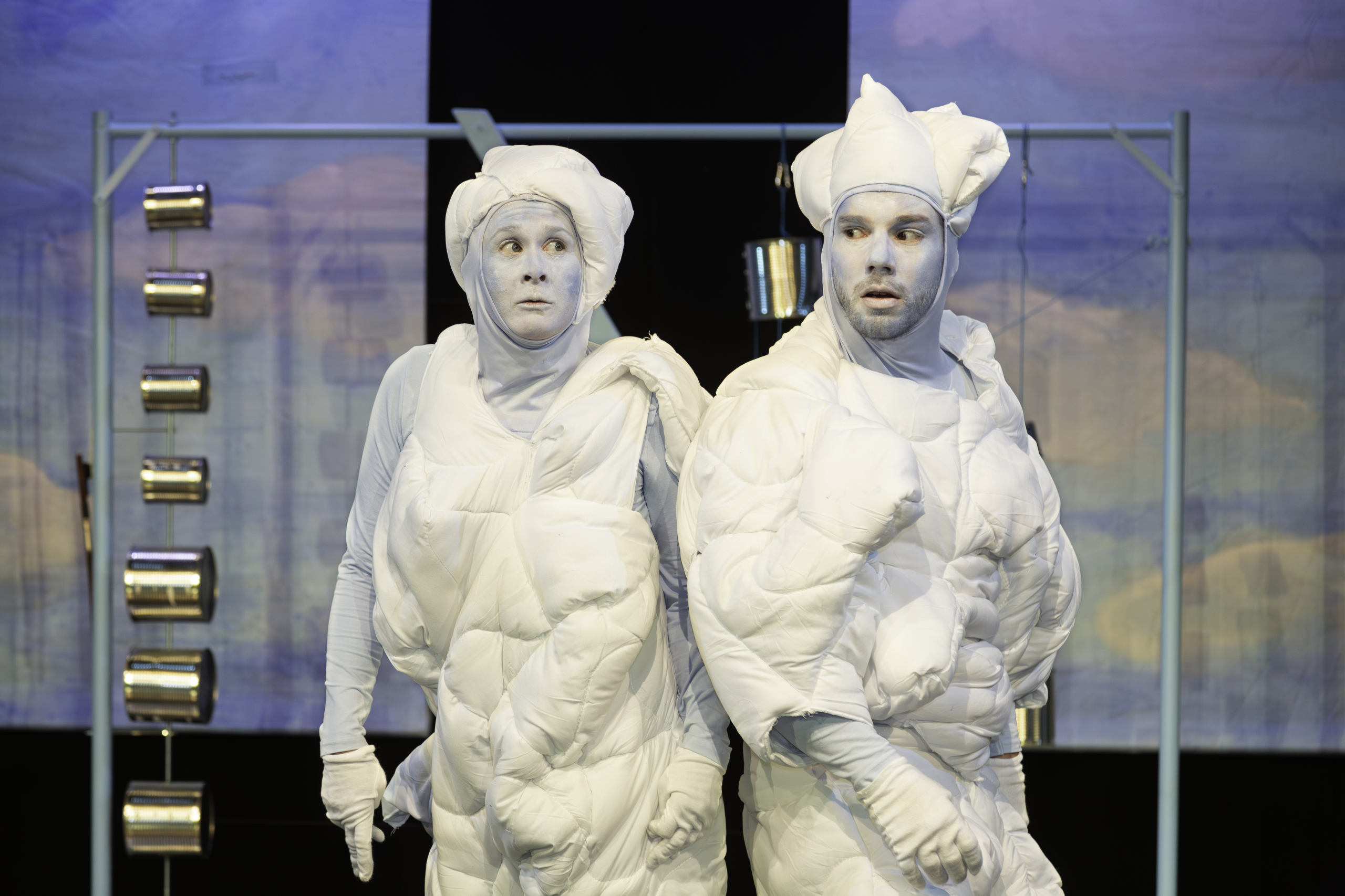 Zwei Personen in weißen, gepolsterten Kostümen mit passenden Mützen und Handschuhen auf einer Bühne mit blauem Hintergrund.