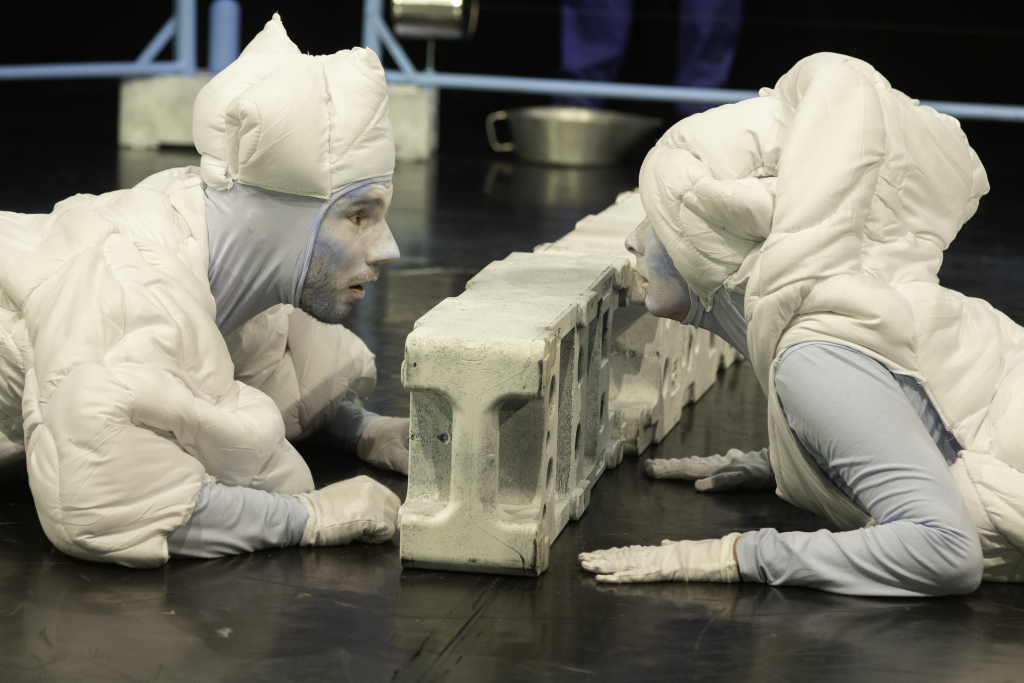 Zwei Personen in weißen, gepolsterten Ganzkörperanzügen liegen sich auf dem Boden gegenüber, getrennt durch eine Reihe grauer Betonblöcke.