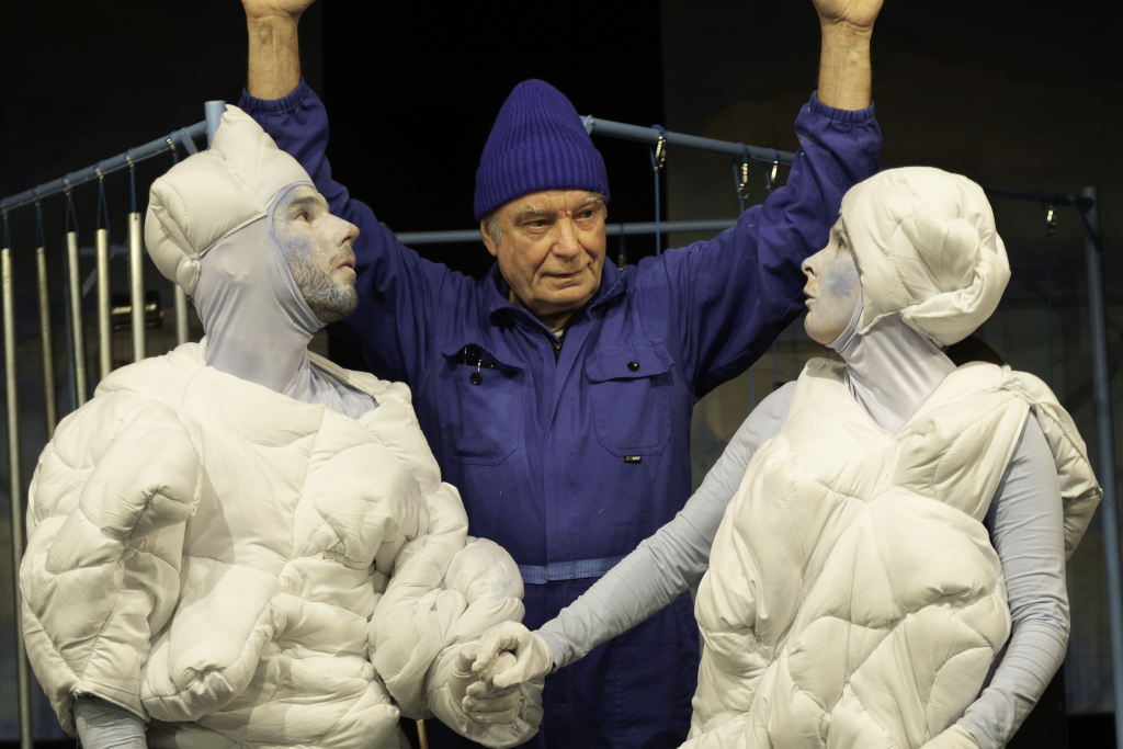 Person in blauer Arbeitskleidung und blauer Mütze steht mit erhobenen Armen zwischen zwei weißen, gepolsterten Schauspieler*innen.