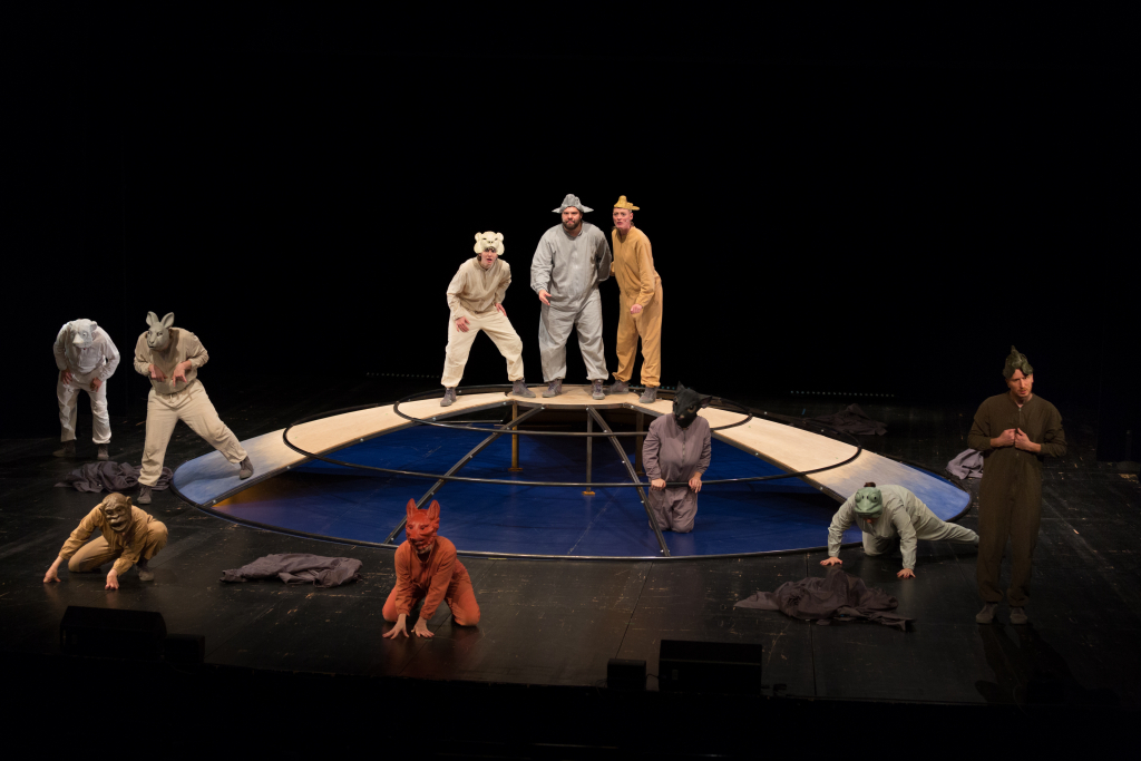 Mehrere Personen in Tierkostümen und -masken stehen und knien um eine kreisförmige Bühne mit blau-weißer Fläche in der Mitte, schwarzer Hintergrund.