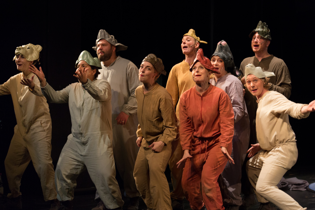 Gruppe von neun Personen in bunten, einfarbigen Kostümen und Tiermützen auf dunkler Bühne, sie gestikulieren lebhaft.