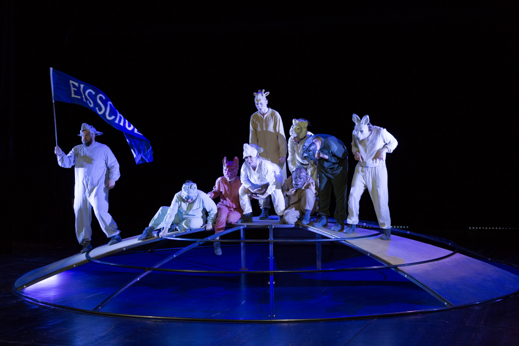 Sieben Personen in Tierkostümen stehen und knien auf einer halbrunden, beleuchteten Bühne, eine Person hält eine blaue Fahne mit dem Schriftzug 'EISSCHOLLE'.