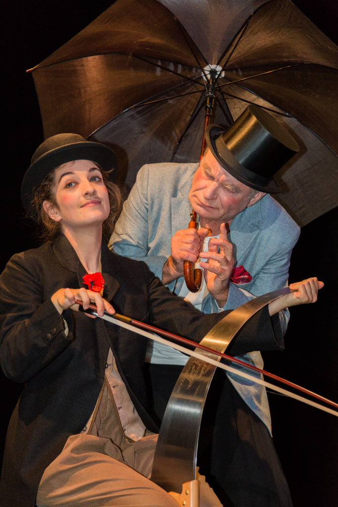 Person mit schwarzem Hut spielt Cello, eine weitere Person hält einen geöffneten Regenschirm über sie.