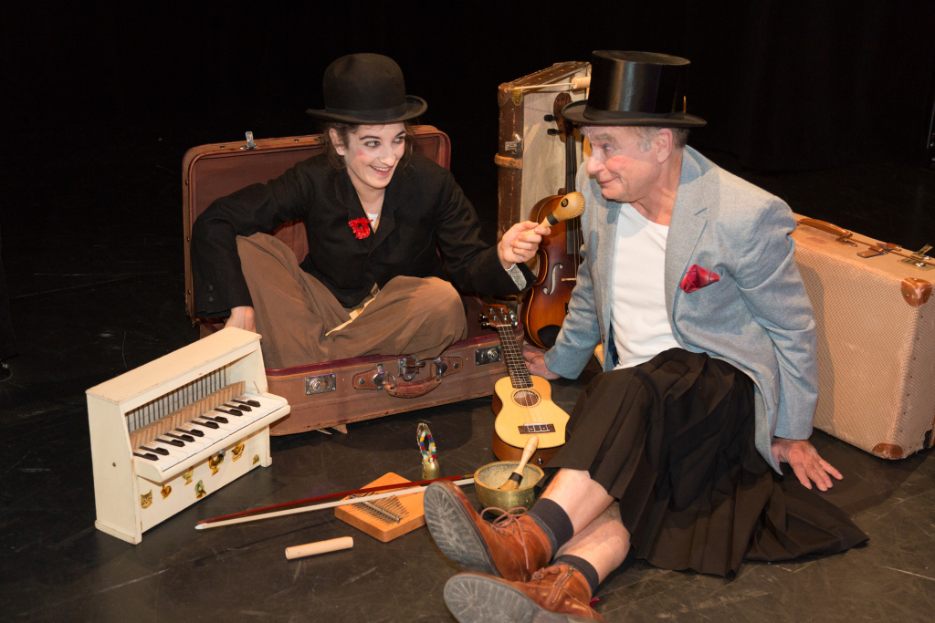 Zwei Personen in Hüten sitzen auf dem Boden, umgeben von Musikinstrumenten und Koffern, eine hält eine kleine Ukulele.