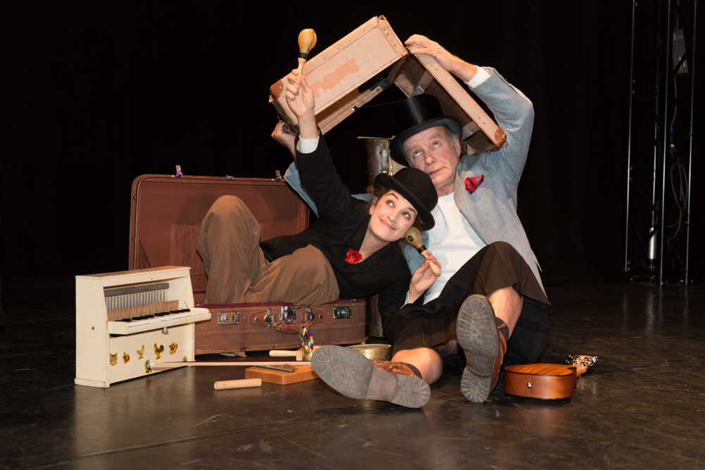 Zwei Personen in Zylinderhüten sitzen auf dem Boden, umgeben von Musikinstrumenten und Koffern, eine hält eine Ukulele, die andere eine Rassel.