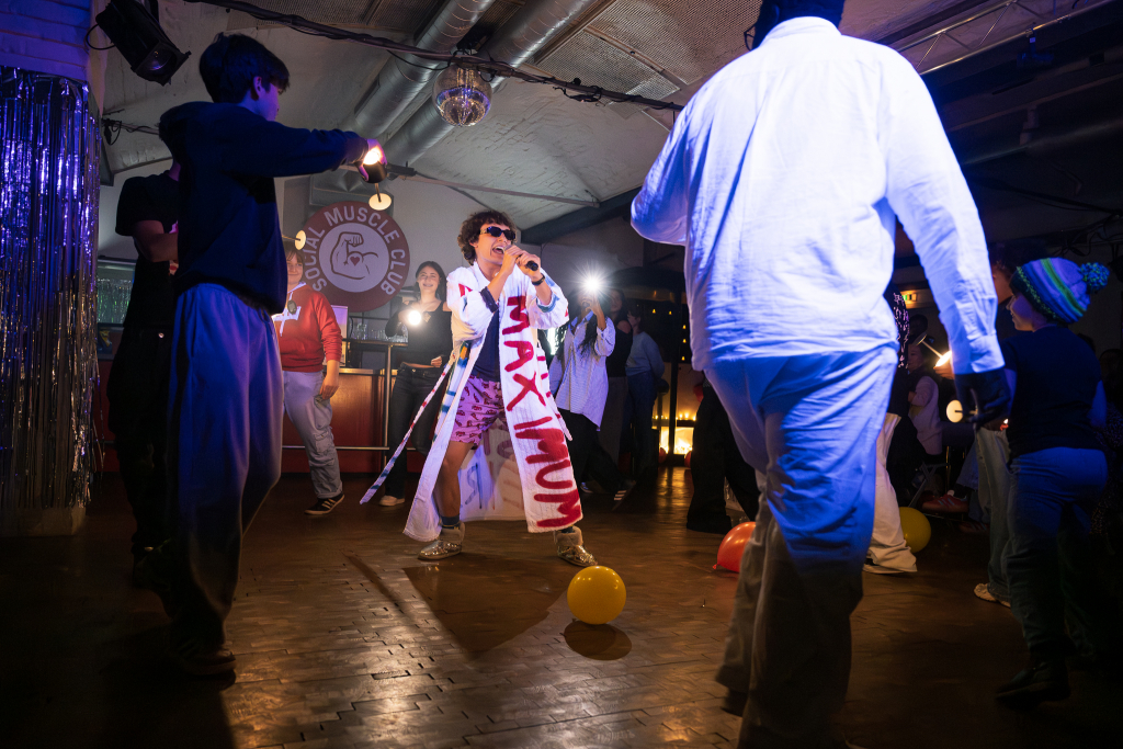 Person mit Sonnenbrille und langem weißen Mantel mit roter Aufschrift singt in einem dunklen Raum vor mehreren tanzenden Personen