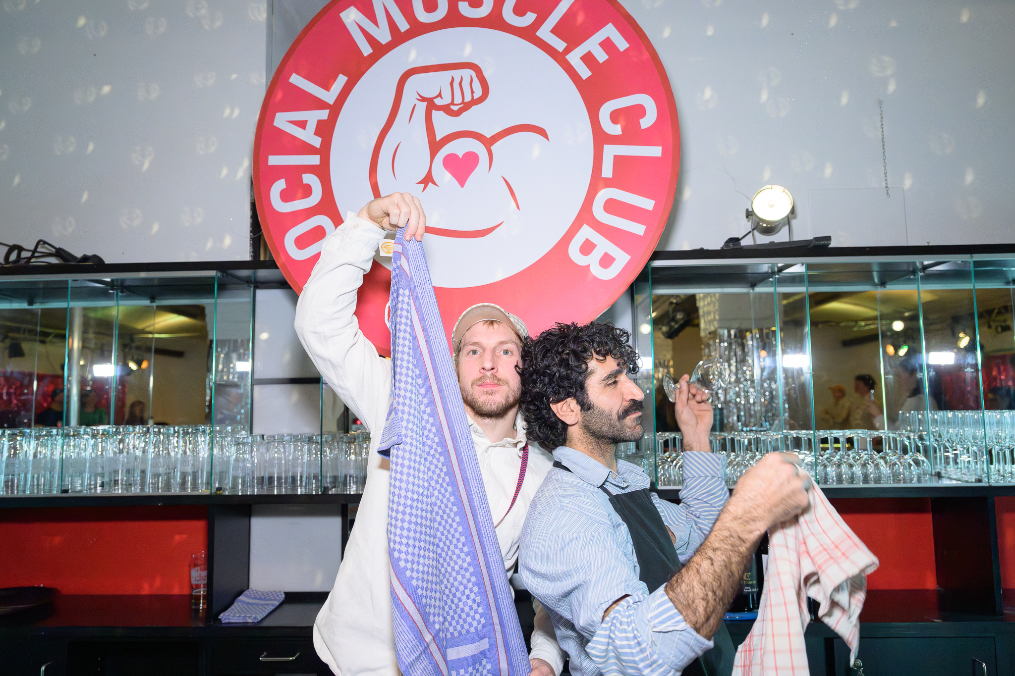 Zwei Männer stehen vor einer Bar mit Gläsern und halten jeweils ein Geschirrtuch hoch, im Hintergrund ein rundes Schild mit der Aufschrift 'SOCIAL MUSCLE CLUB' und einem muskulösen Arm mit Herzsymbol