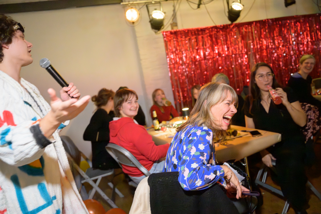 Person mit Mikrofon spricht vor einer Gruppe sitzender Menschen in einem Raum mit rotem Glitzervorhang