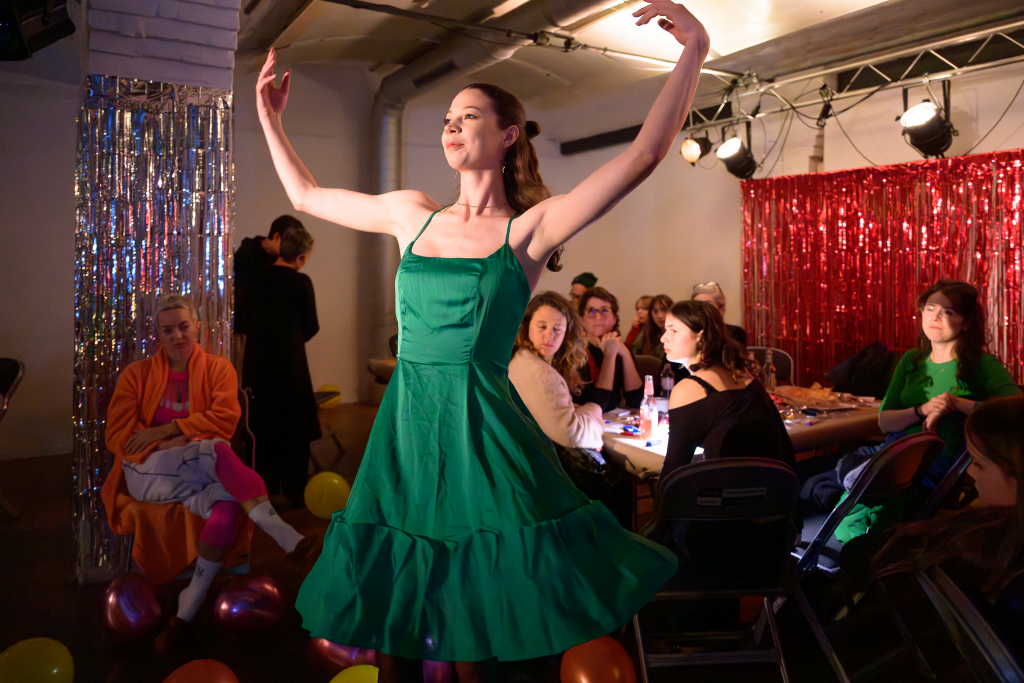 Person mit Mikrofon spricht vor einer Gruppe sitzender Menschen in einem Raum mit rotem Glitzervorhang