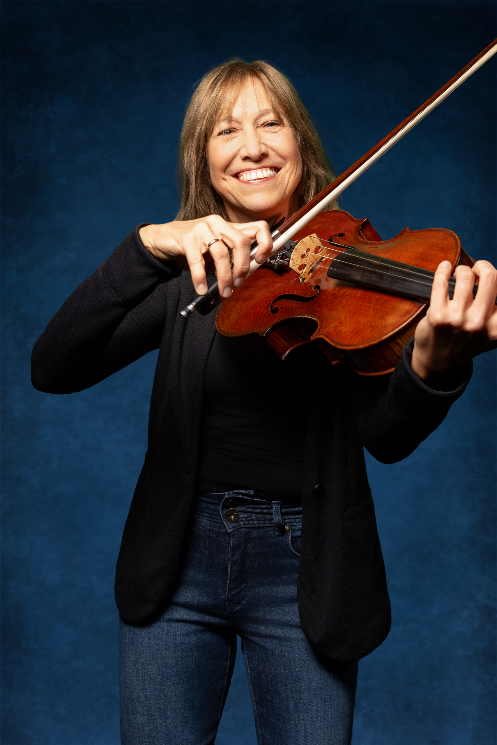 Lächelnde Frau mit Geige in der Hand, steht vor blauem Hintergrund, trägt schwarze Jacke und Jeans.
