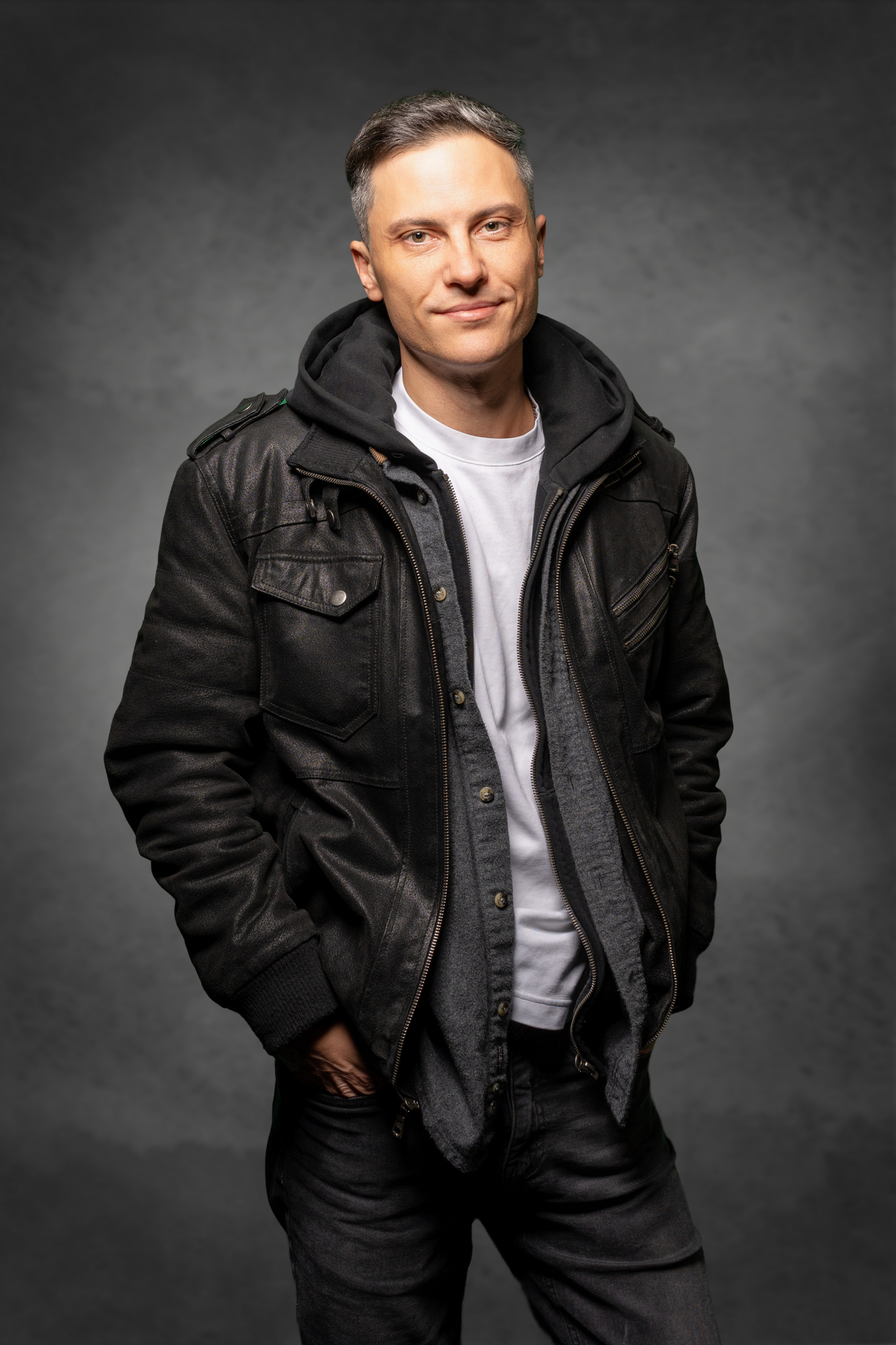 Porträt eines Mannes in einer schwarzen Lederjacke mit Kapuze, lächelnd vor grauem Hintergrund.