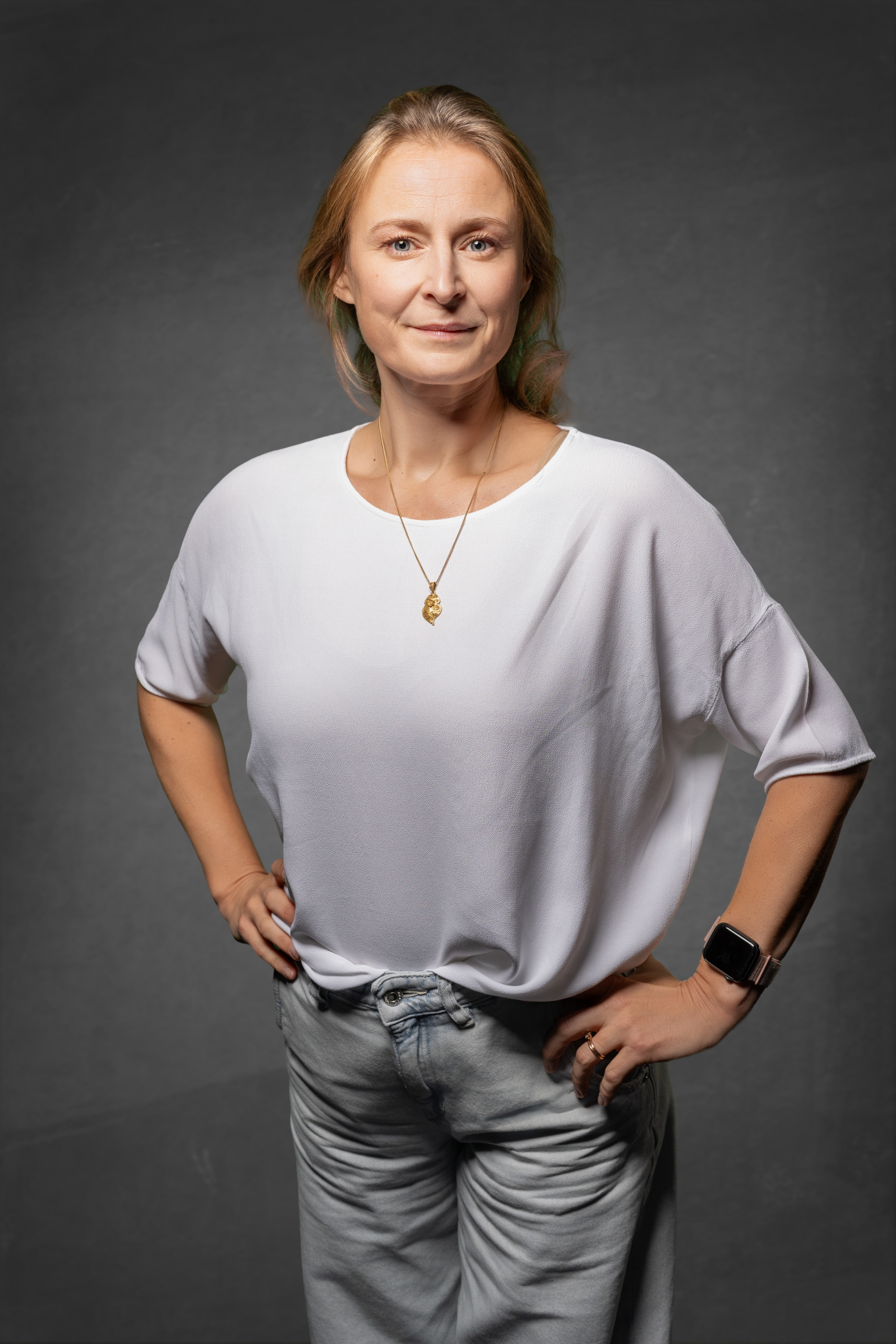 Person mit blonden Haaren in weißem Oberteil und grauen Hosen, lächelnd vor dunklem Hintergrund.