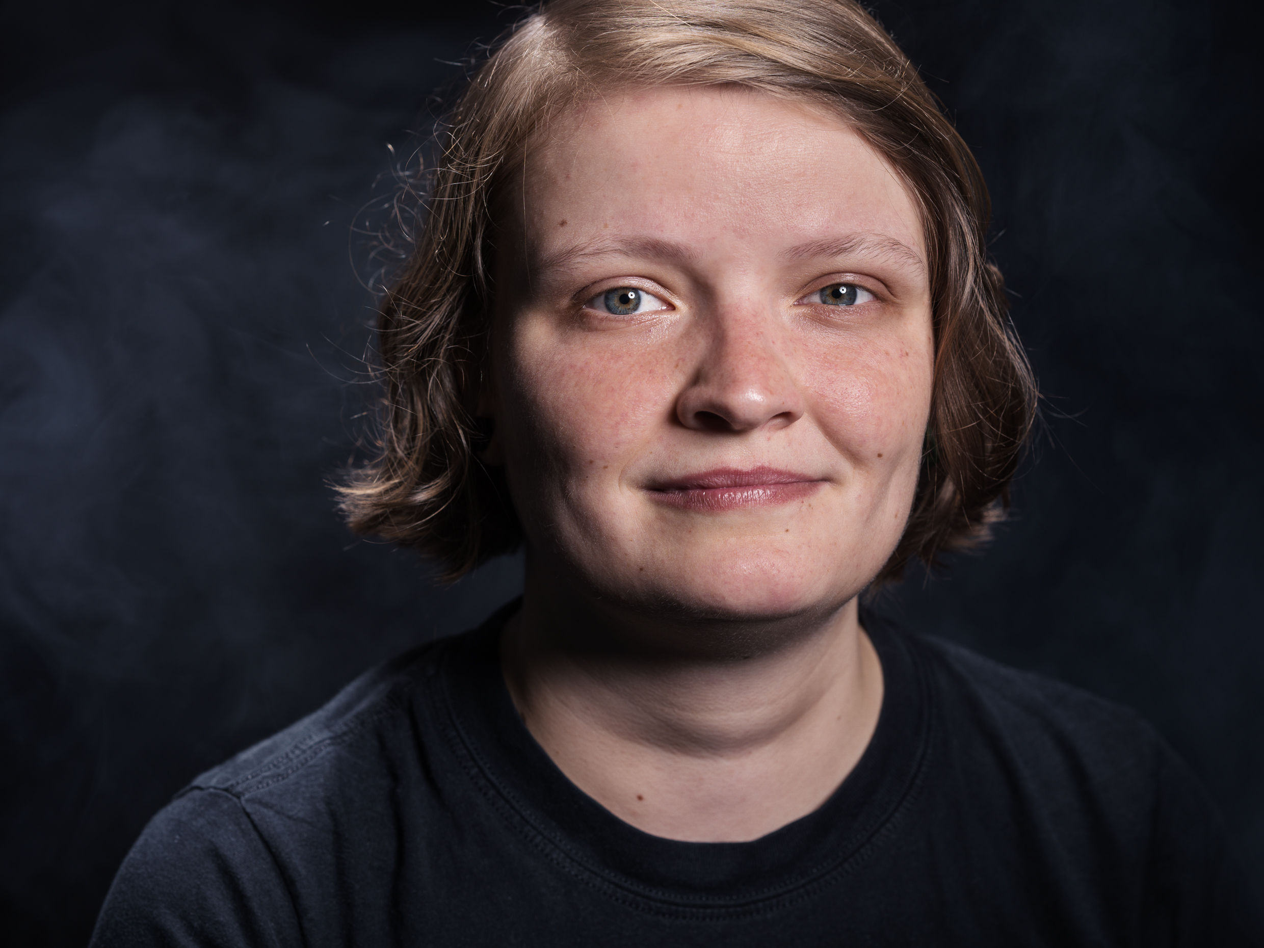 Portrait einer Person mit kurzen Haaren und einem freundlichen Gesichtsausdruck auf dunklem Hintergrund.