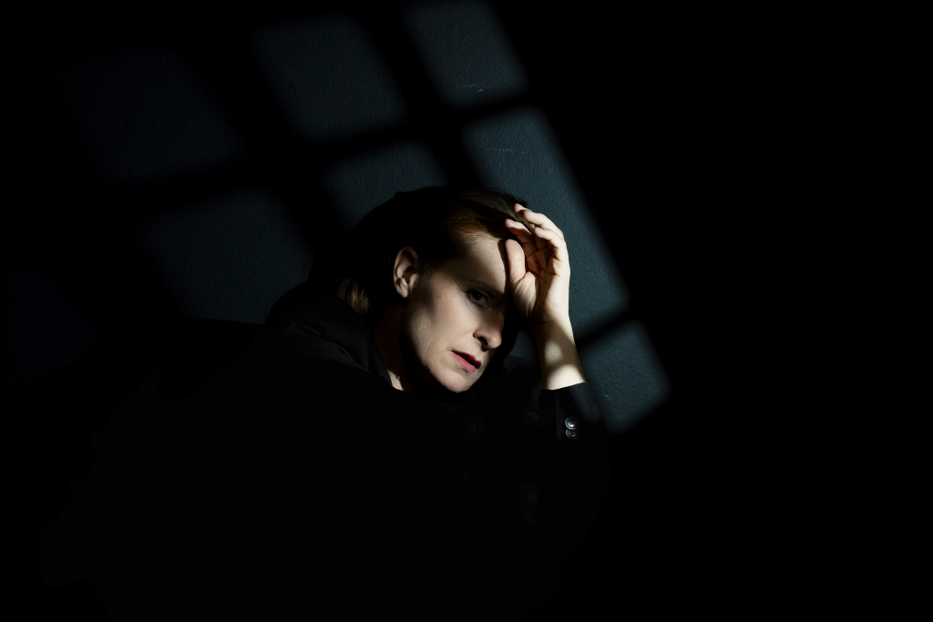 Person hebt eine Hand an den Kopf vor einem dunklen Hintergrund mit einem Fensterkreuz aus Licht