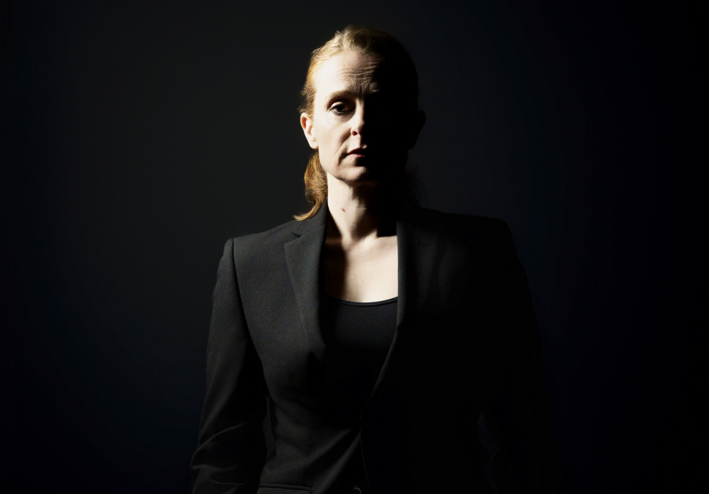 Siluette einer Frau in einem schwarzen Anzug mit zurückgebundenen Haaren vor dunklem Hintergrund.