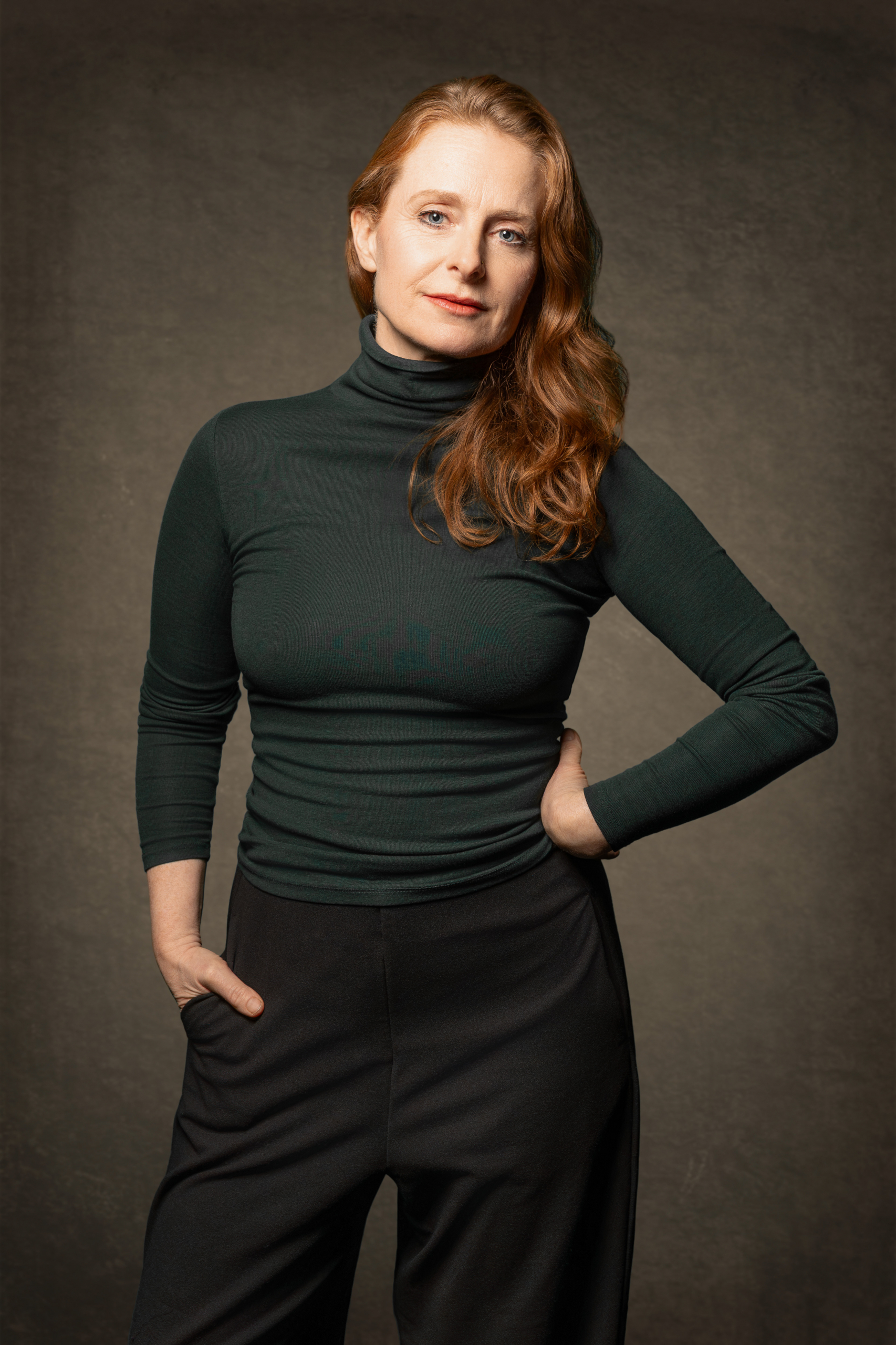 Frau mit langen, gewellten Haaren trägt schwarzen Rollkragenpullover und schwarze Hose, steht mit einer Hand in der Hosentasche und der anderen am Hüftgelenk vor neutralem Hintergrund