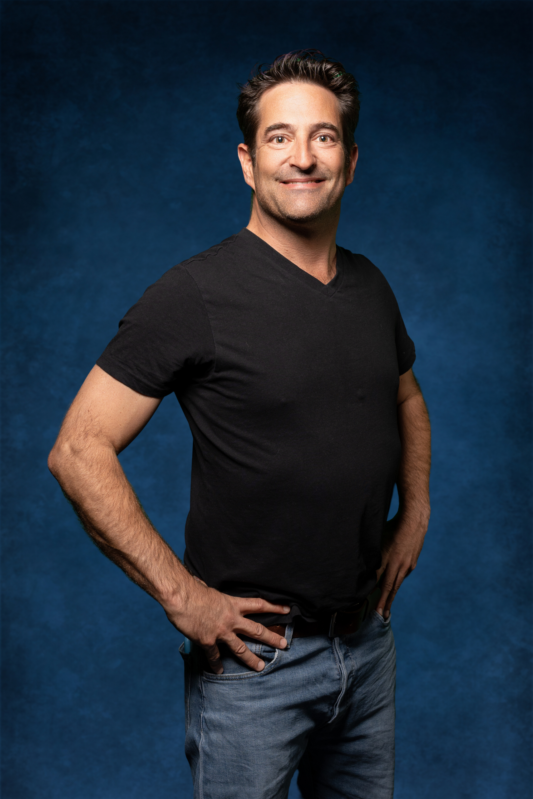 Portrait eines lächelnden Mannes in schwarzem T-Shirt und Jeans vor blauem Hintergrund.