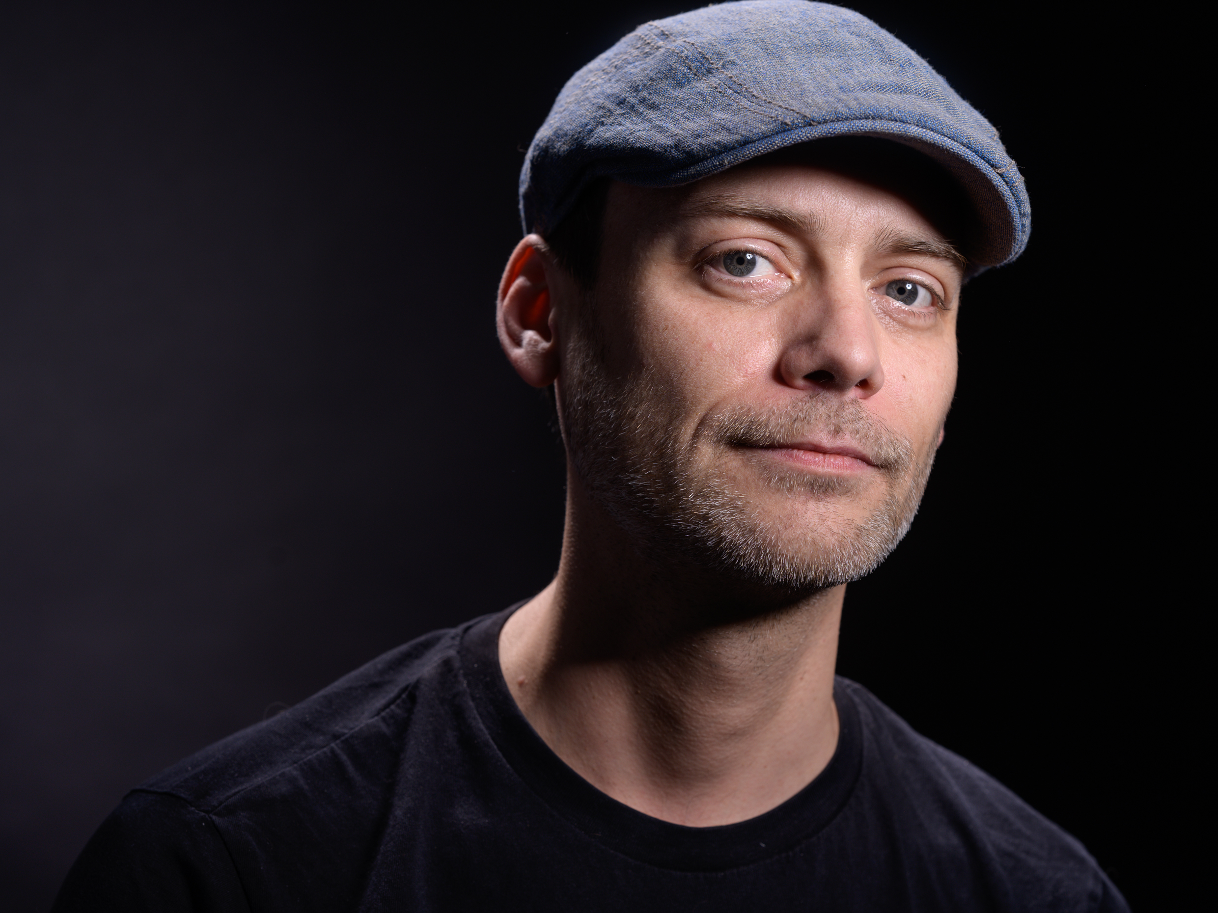 Mann mit blauer Schiebermütze und schwarzem T-Shirt vor dunklem Hintergrund.